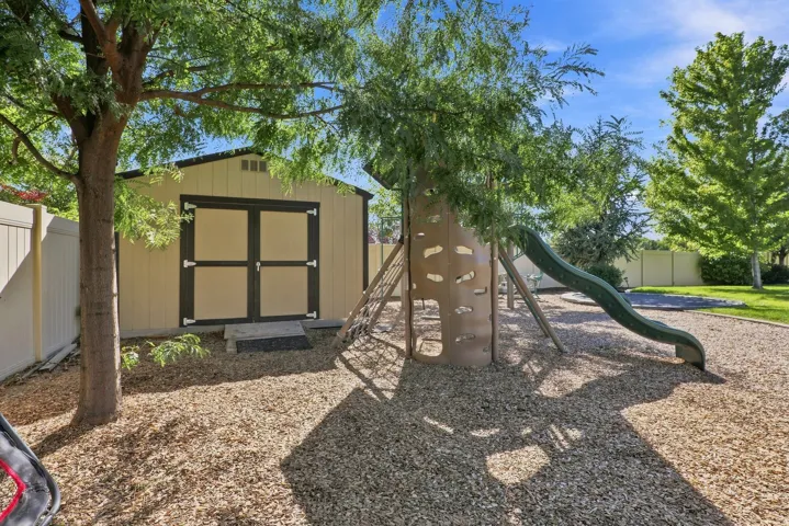 Enclosed outdoor area featuring a storage shed with double doors, wood chip ground cover, a play structure with a climbing wall, and a slide