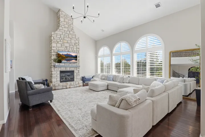 Vaulted ceiling with an elegant chandelier, stone-clad fireplace with a hearth, three arched windows with plantation shutters, dark wood-finish flooring