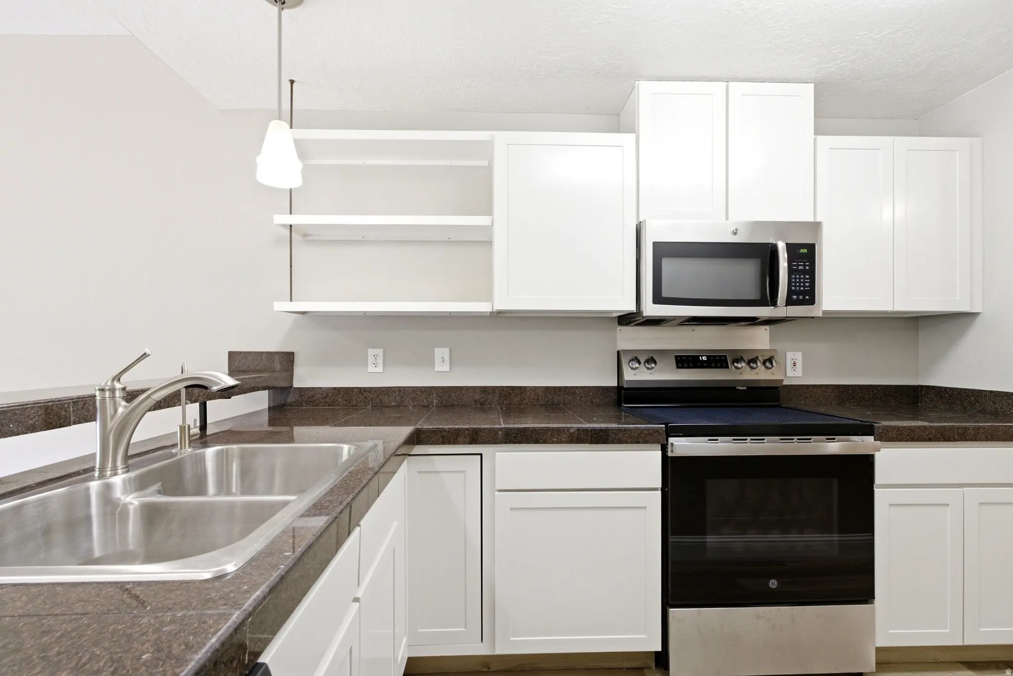 Kitchen featuring white cabinetry with brushed nickel hardware, dark stone-finish countertops, stainless steel double basin sink with brushed nickel faucet, stainless steel overhead microwave, and stainless steel range with a black cooktop