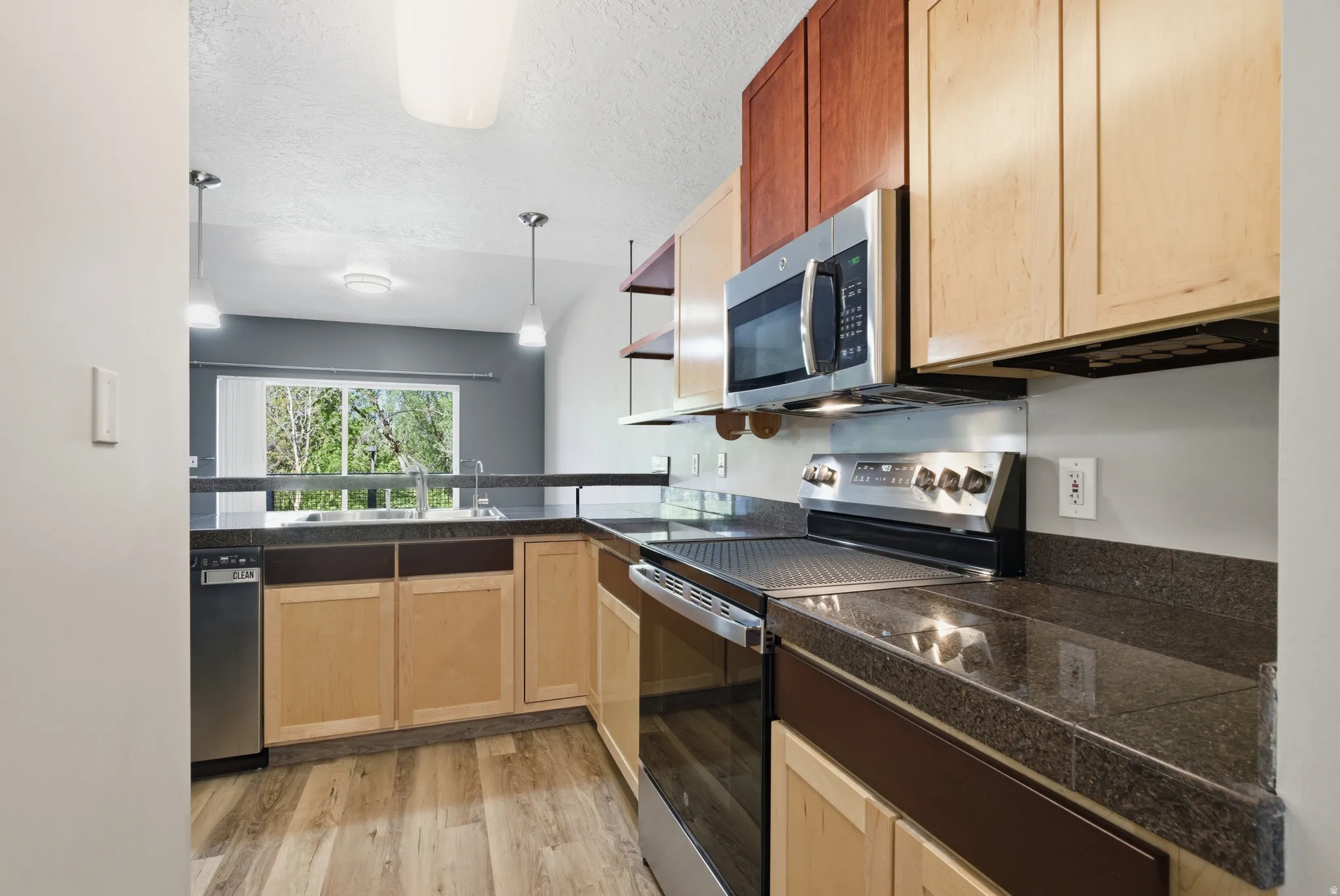Kitchen featuring tile countertops, stainless steel appliances, light wood finish cabinetry, light wood-style floors, and a textured ceiling