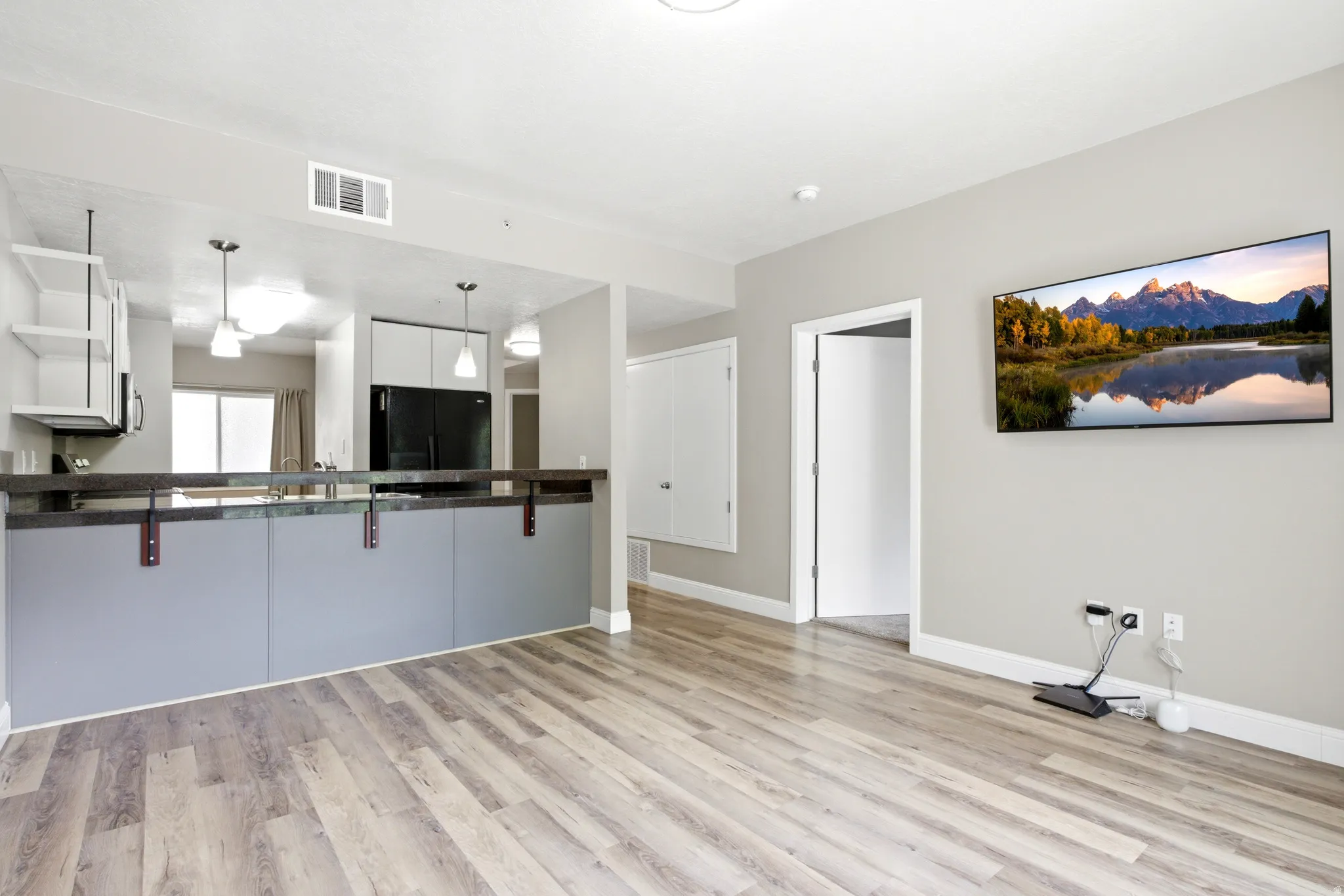 Open-concept living space featuring wood-finish flooring, a kitchen island with a dark countertop and grey paneling, white shaker-style upper cabinets, and black appliances