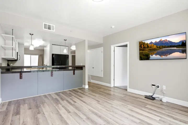 Open-concept living space featuring wood-finish flooring, a kitchen island with a dark countertop and grey paneling, white shaker-style upper cabinets, and black appliances