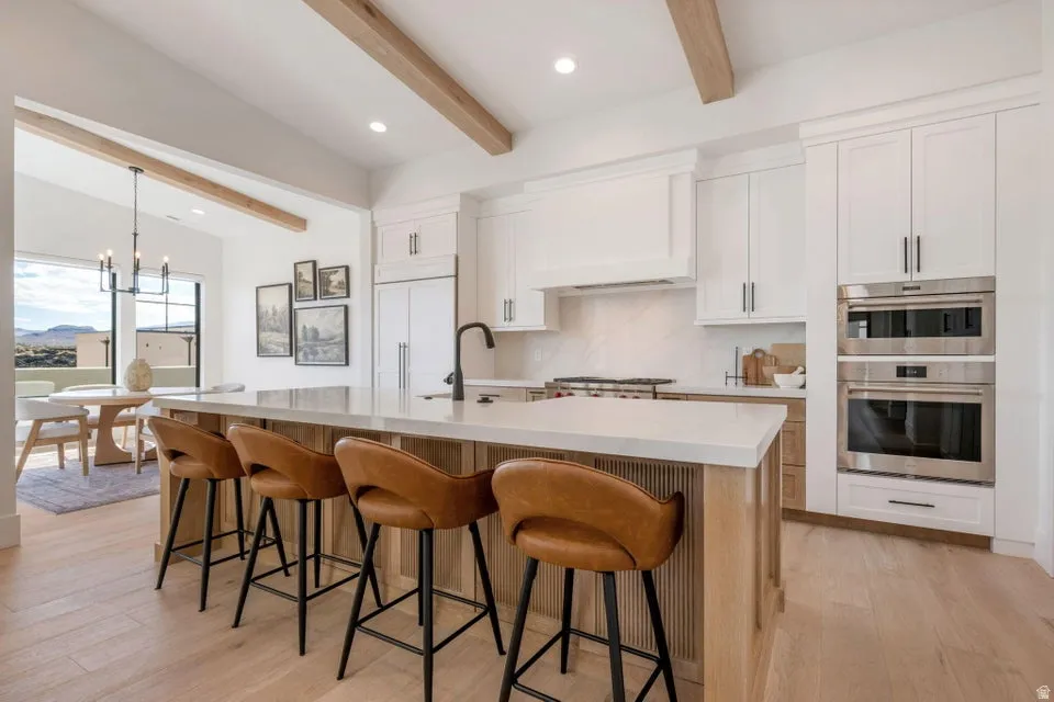 Kitchen with light wood-style flooring, beamed ceiling, two tone color scheme, a breakfast bar, and decorative backsplash