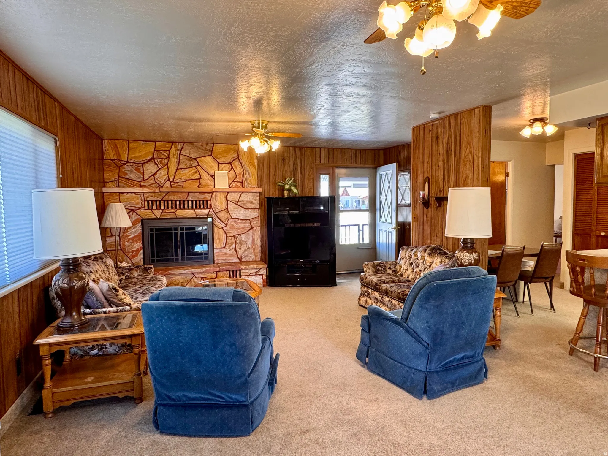 Carpeted living room with wood walls, ceiling fan, a stone fireplace, and a textured ceiling
