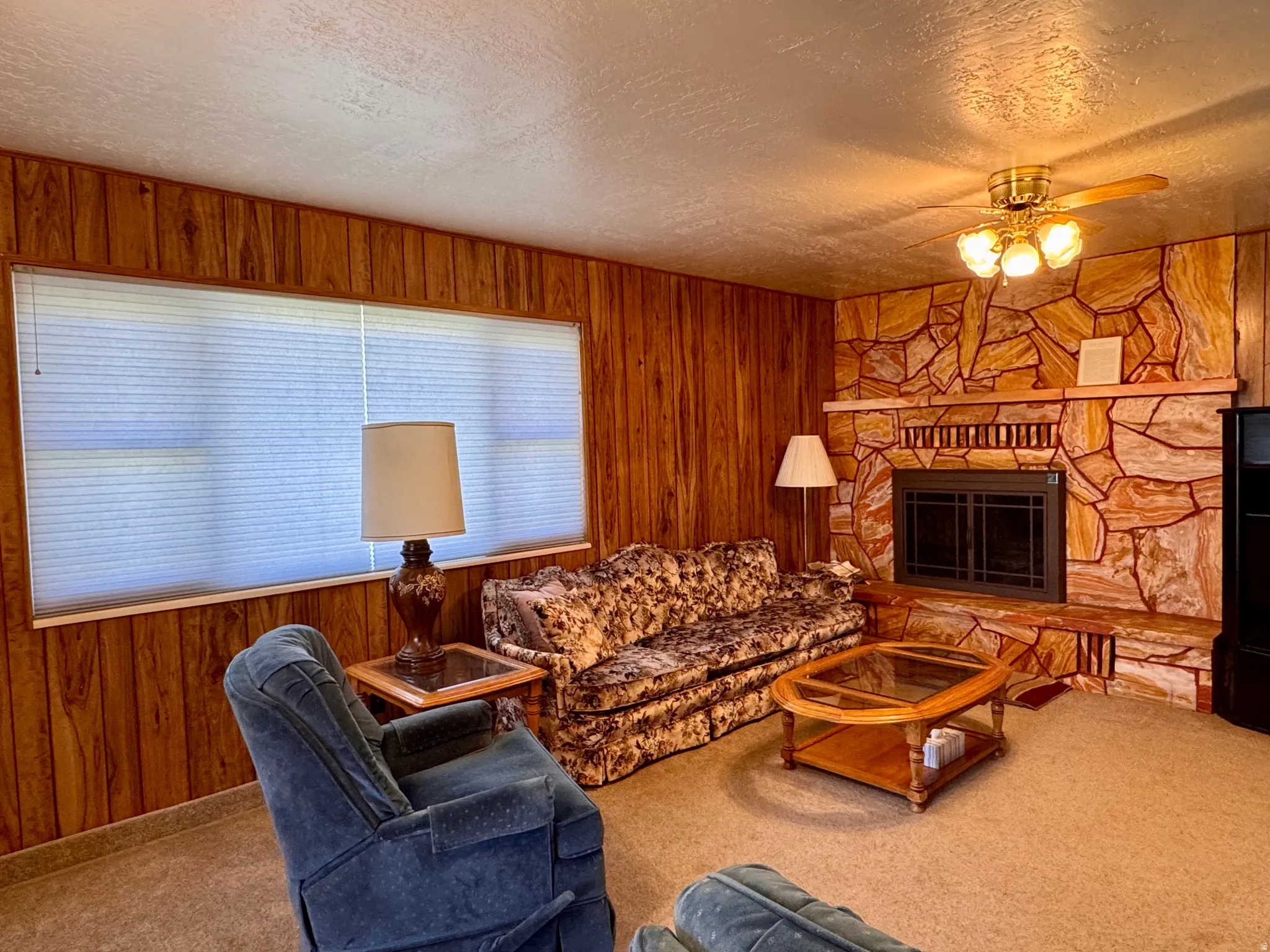 Living room featuring wood walls, a textured ceiling, a ceiling fan, carpet floors, and a stone fireplace