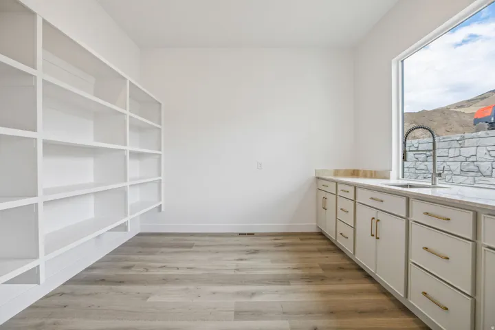 Extensive built-in shelving, wood-finish flooring, white cabinetry with gold-tone hardware, undermount sink with a gooseneck faucet, and a large window