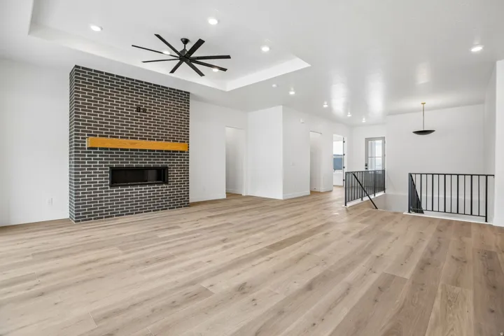 Spacious room featuring wood-finish flooring, a contemporary fireplace with a black brick surround and wood mantel, and a tray ceiling with recessed lighting and a ceiling fan