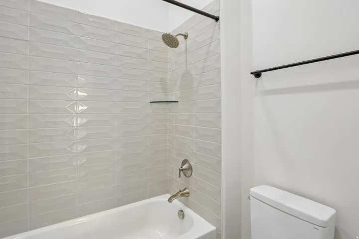 Bathtub with textured tile surround, brushed nickel fixtures, a glass shower shelf, and a matte black towel bar