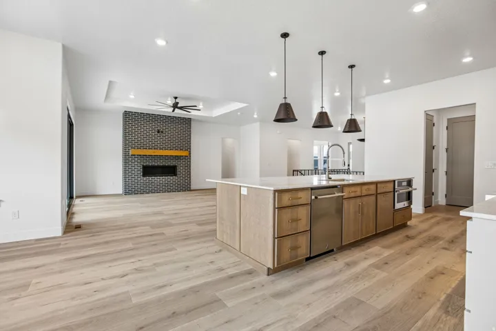 Wood-finish flooring throughout, kitchen island with built-in stainless-steel appliances, recessed lighting, fireplace with a brick surround and wood mantel, and tray ceiling with a ceiling fan