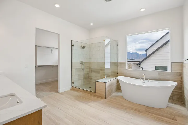 Freestanding soaking tub, glass-enclosed shower with tiled walls, wood-finish flooring, and a single vanity with stone countertop