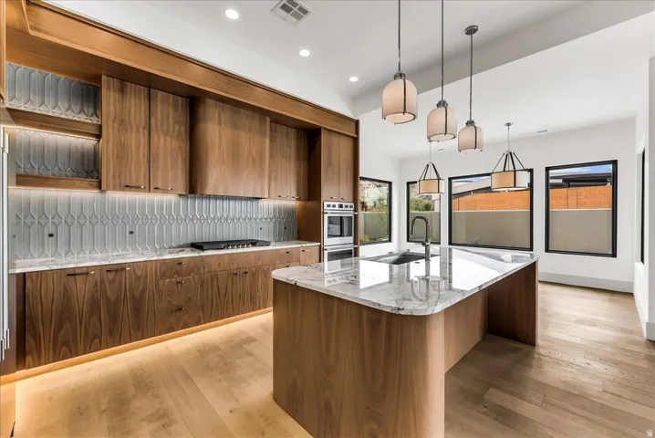 Gourmet kitchen featuring wood-finish cabinetry, a large kitchen island with a stone countertop and integrated sink, a tiled backsplash, wood-finish flooring, and pendant lighting