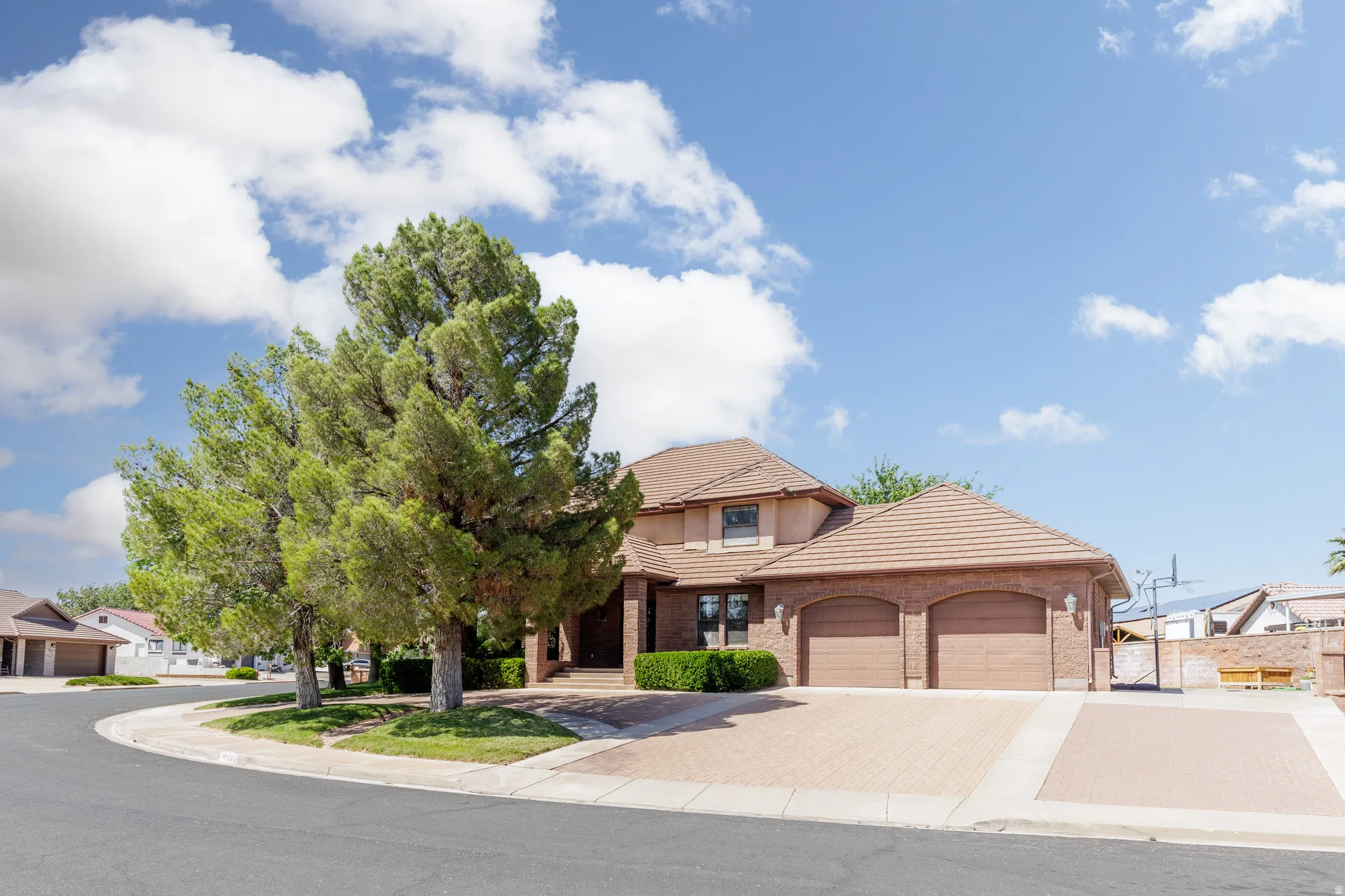 Brick exterior with a tile roof, featuring a two-car garage, mature trees, and a paver driveway