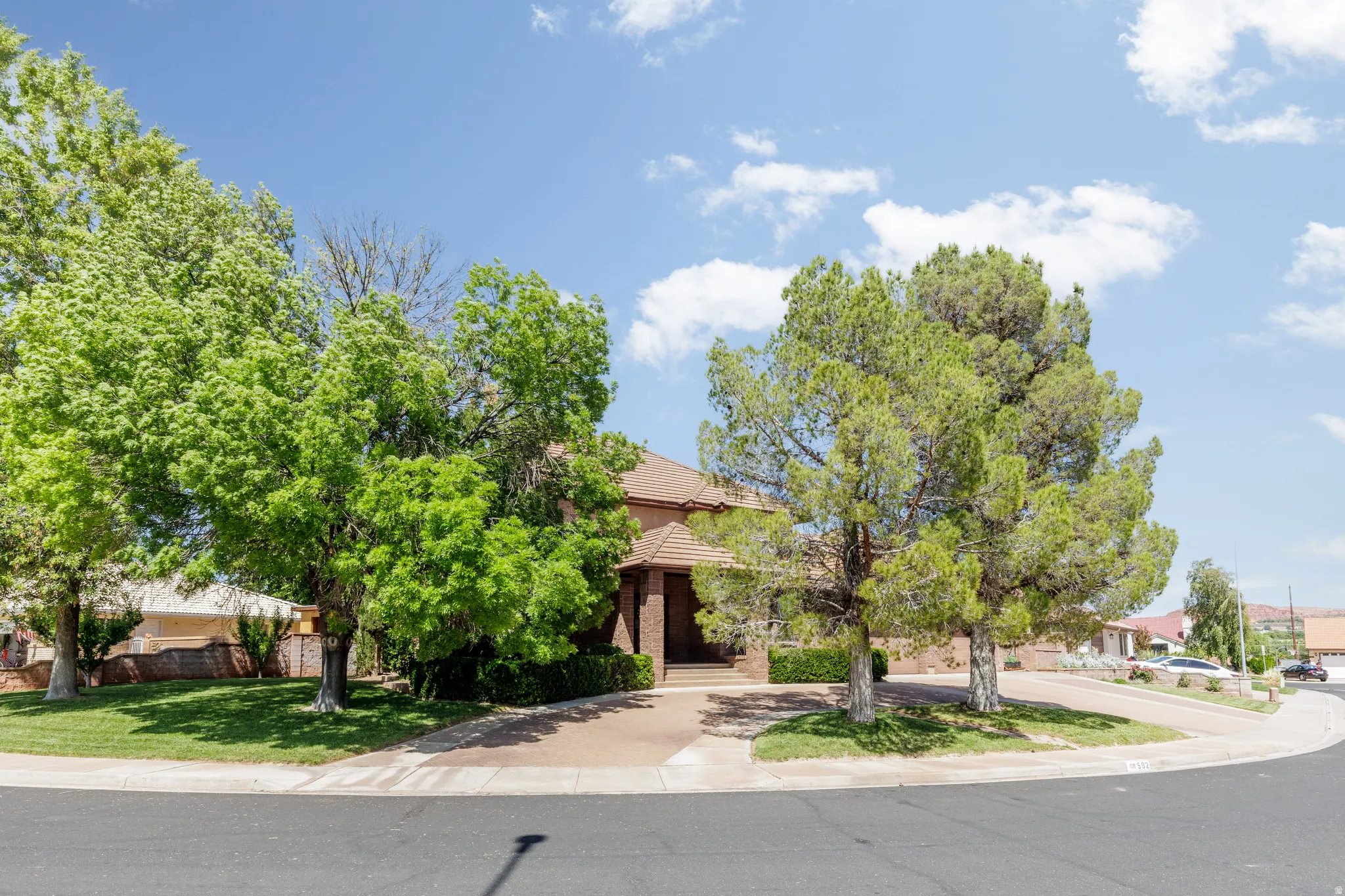 Brick exterior residence with a tile roof, mature trees, a driveway, and a manicured lawn