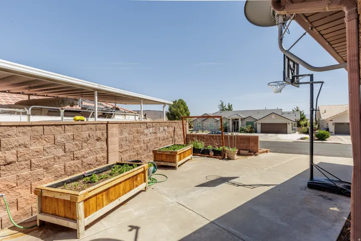 Paved outdoor area featuring raised garden beds, a block retaining wall, and an attached covered patio structure