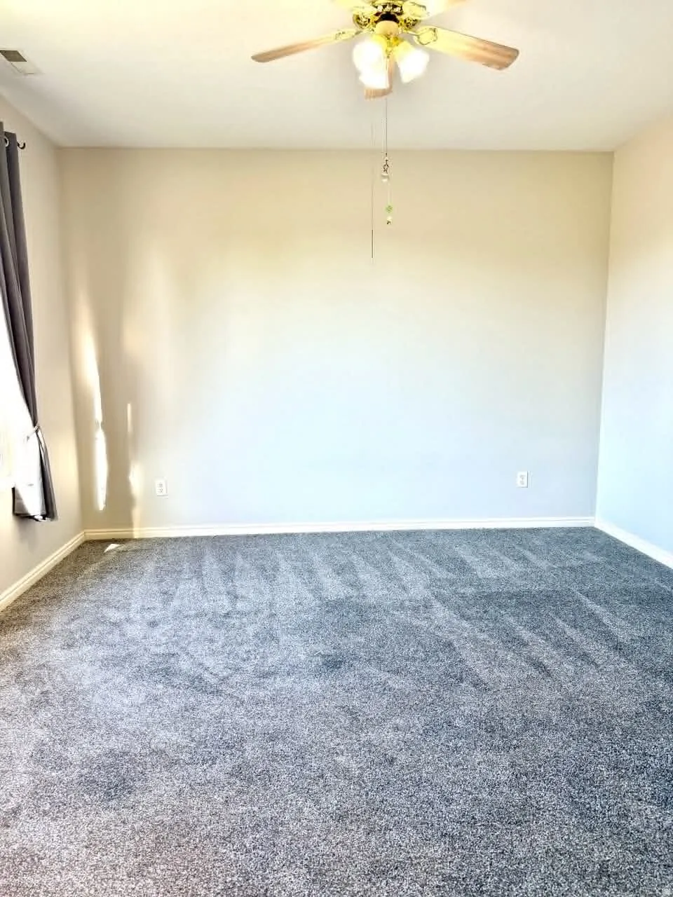Spacious interior room featuring light-colored walls, gray carpeting, a ceiling fan with integrated lighting, and a window with dark drapery