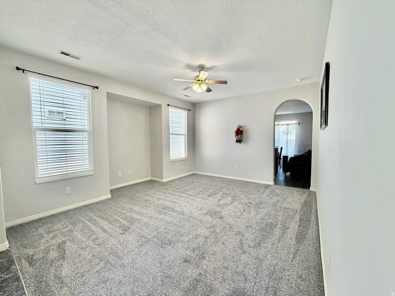 Spacious room featuring light gray carpet flooring, multiple windows with blinds, a ceiling fan with light fixture, an arched doorway, and neutral wall paint