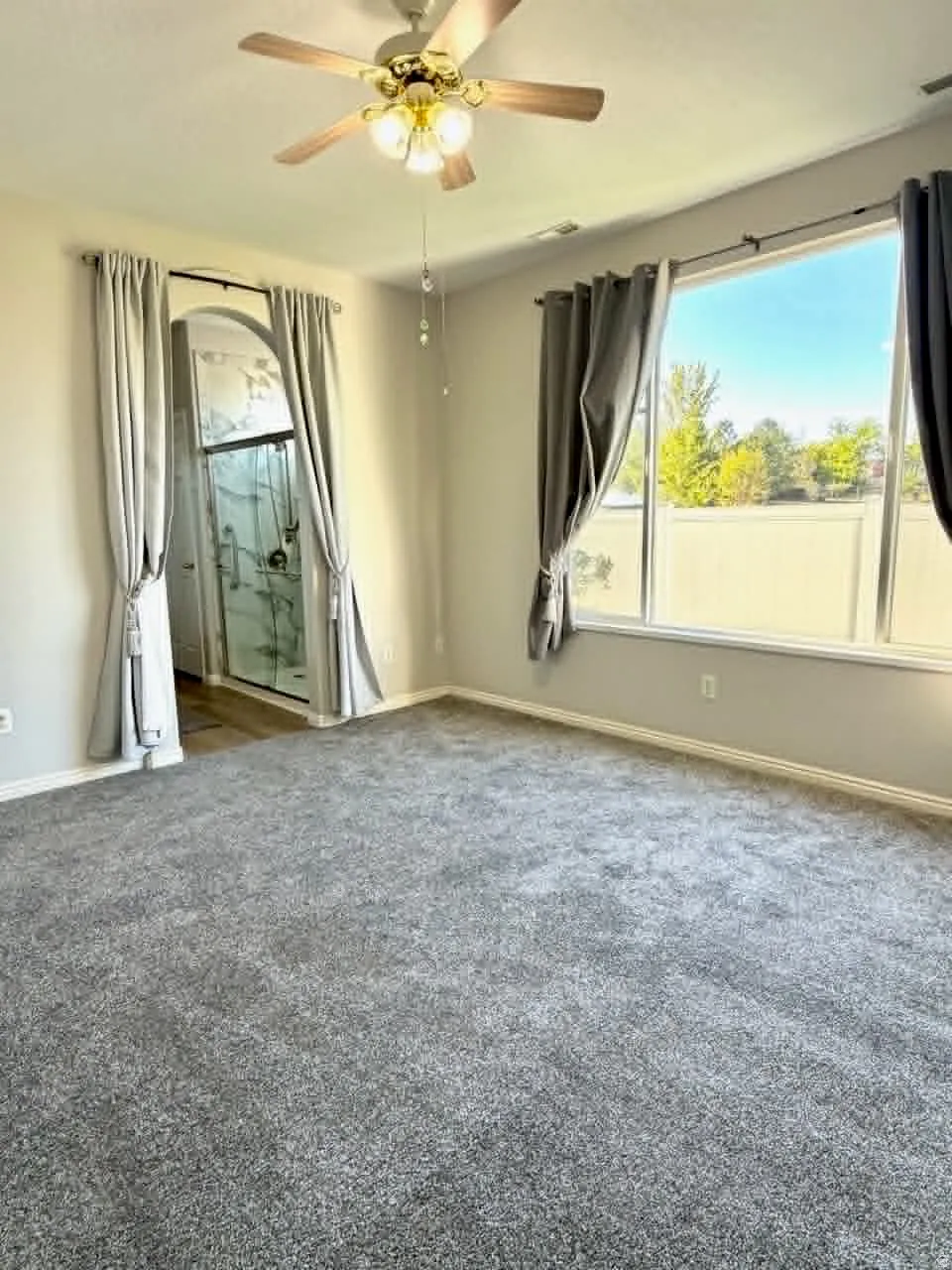 Spacious carpeted room featuring a ceiling fan, large windows, and an arched entry to an en-suite bathroom with a glass-enclosed shower