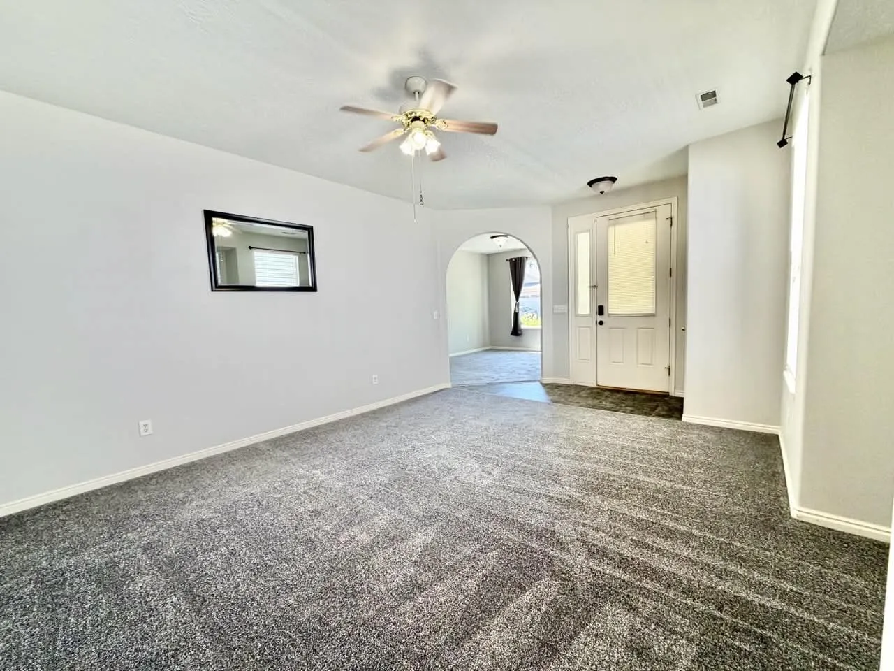 Spacious living area featuring new gray carpet, white painted walls, a ceiling fan with light fixture, and a front entry door with a frosted glass panel and sidelight