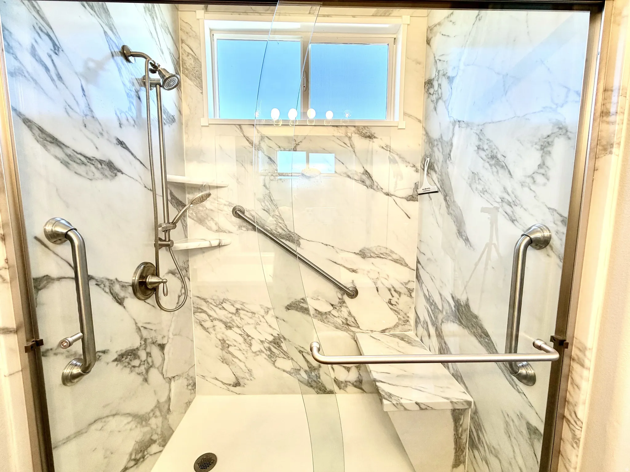 Walk-in shower featuring marble-patterned wall panels, a built-in shower bench, brushed nickel fixtures, glass enclosure, and integrated grab bars