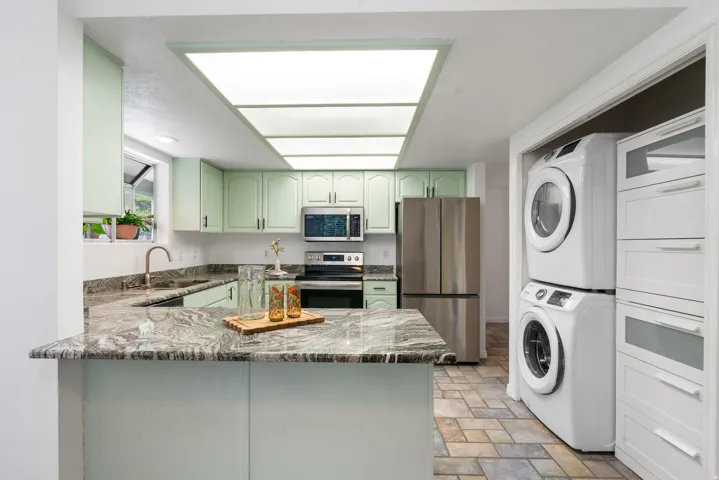 Modern kitchen and laundry suite featuring soft-green cabinetry, stainless steel appliances, granite-style countertops, skylight illumination, and stacked washer-dryer units with white cabinetry