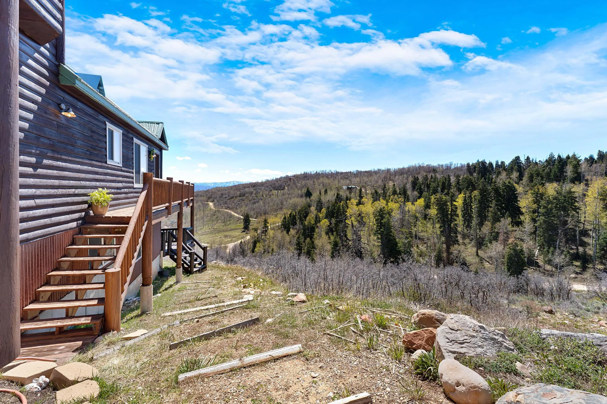 Log cabin exterior featuring a multi-level deck, wood stairs, and natural landscaping with mature trees and distant hills