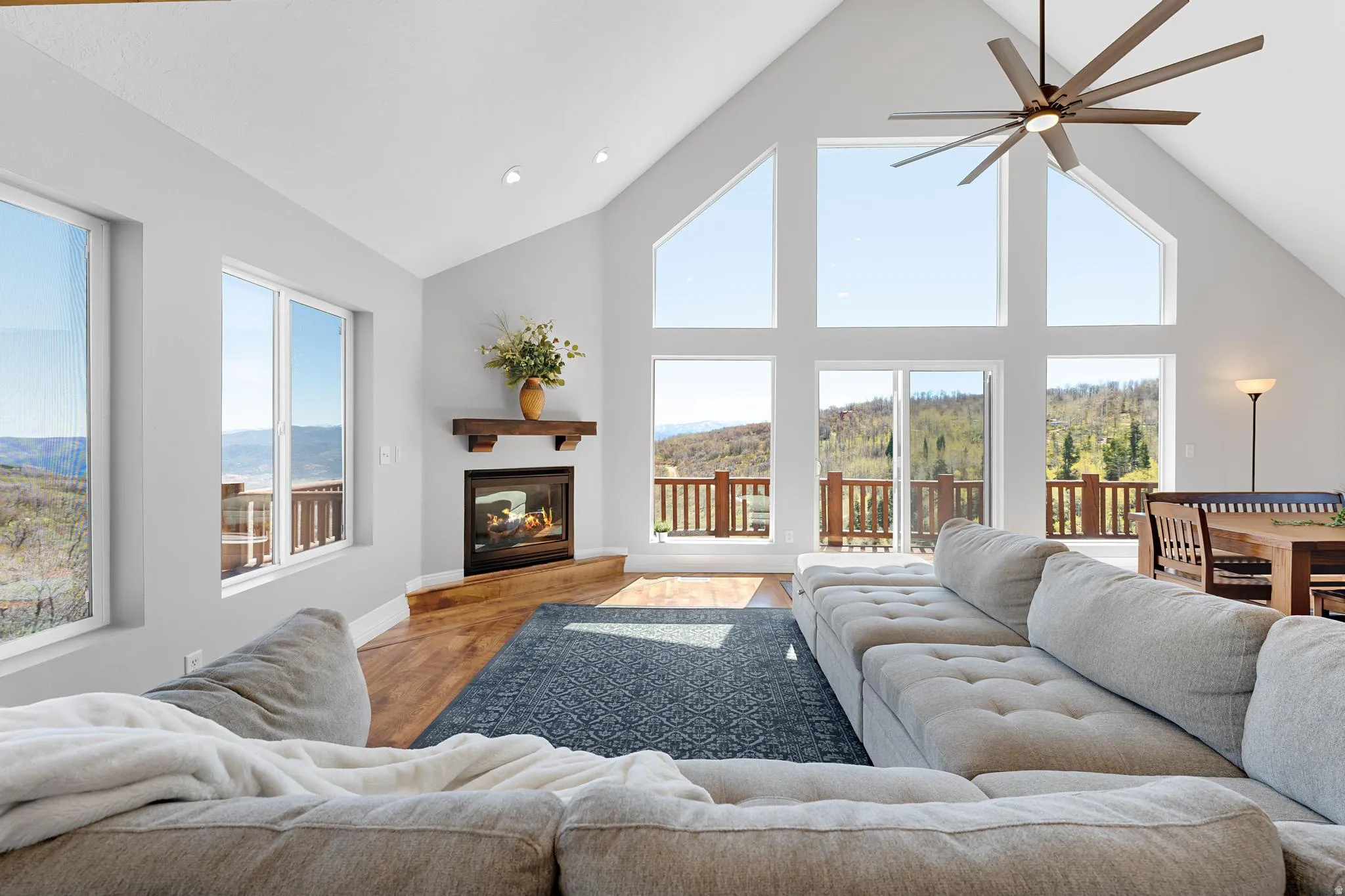 Spacious living area featuring high vaulted ceilings, expansive window walls, a built-in fireplace with a wood mantle, wood-finish flooring, and direct access to an outdoor deck