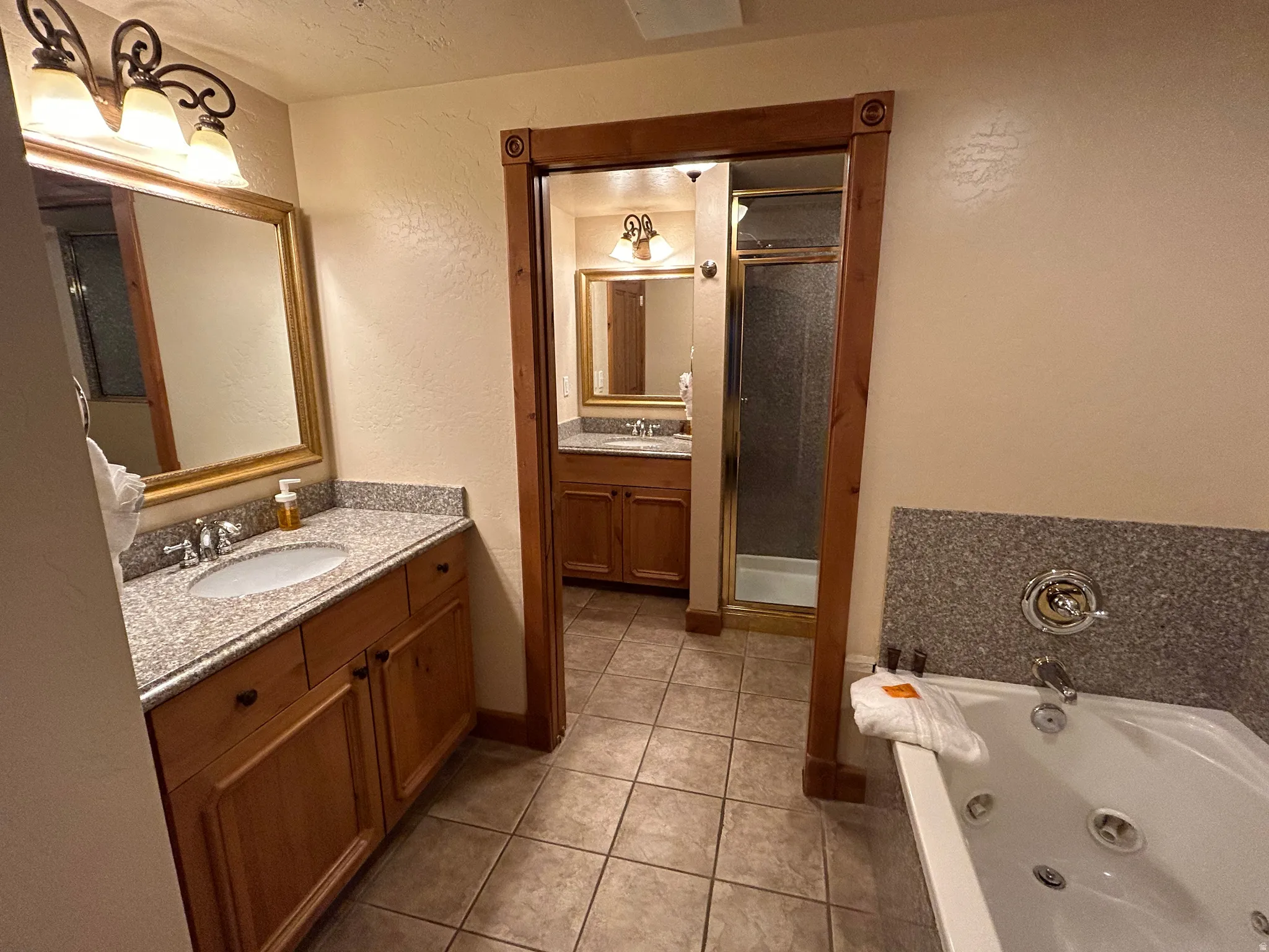 Bathroom featuring a textured wall, two vanities, a shower stall, light tile patterned floors, and a jetted tub