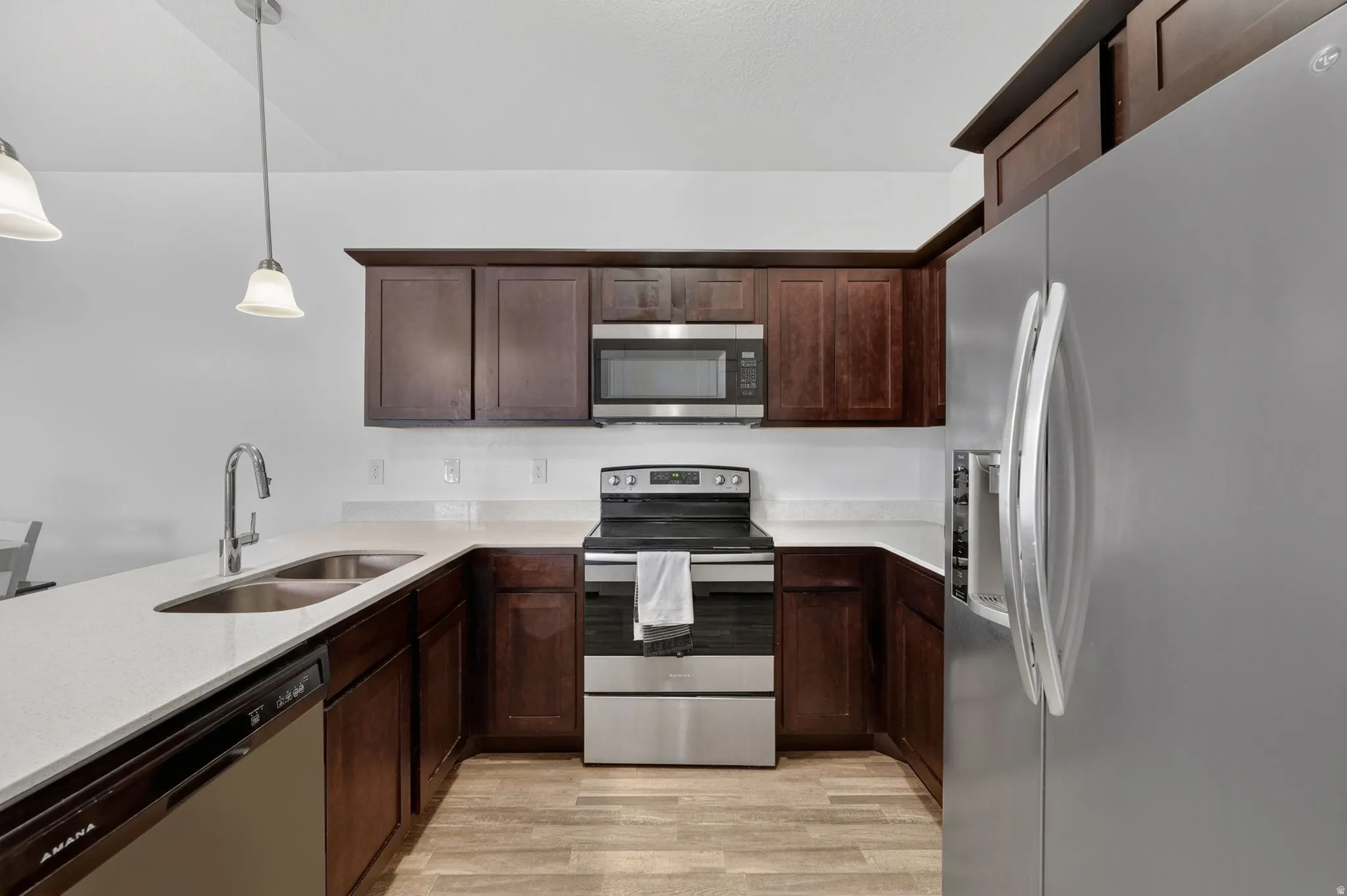 Kitchen featuring stainless steel appliances, dark wood finish cabinetry, decorative light fixtures, light stone counters, and light wood-type flooring