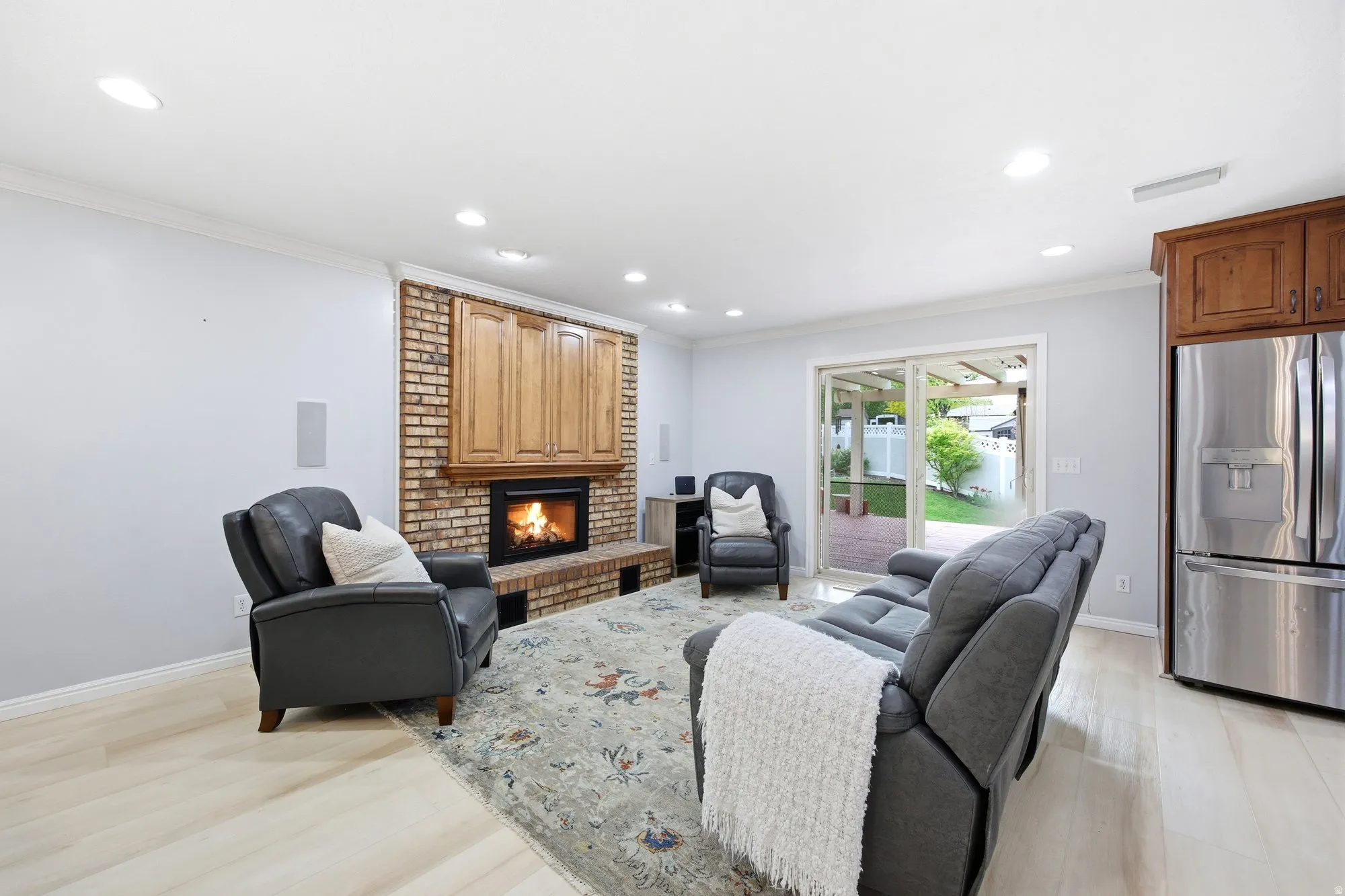 Cozy living  room featuring a brick fireplace, television cabinet,  ornamental molding, light wood  flooring, slider to back deck and recessed lighting