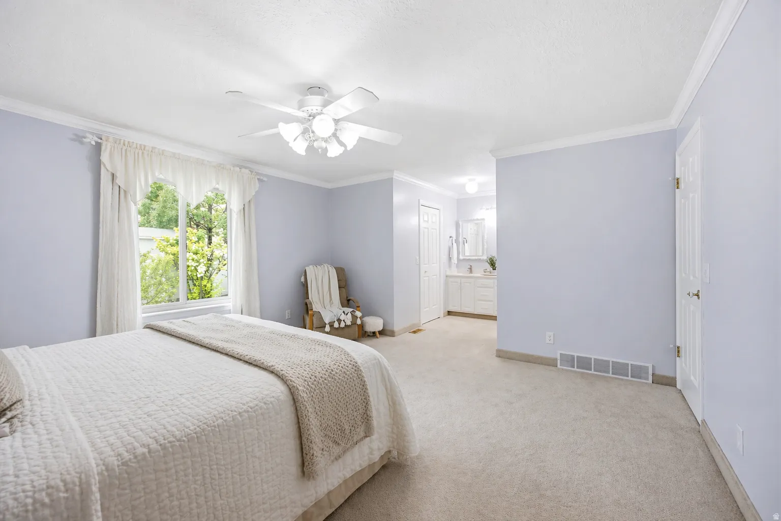 Huge primary bedroom with ornamental molding, light colored carpet, lots of natural light and a ceiling fan