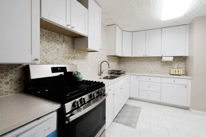 White cabinetry with brushed nickel hardware, stainless steel gas range, light-toned countertop, hexagon tile backsplash, and light tile flooring