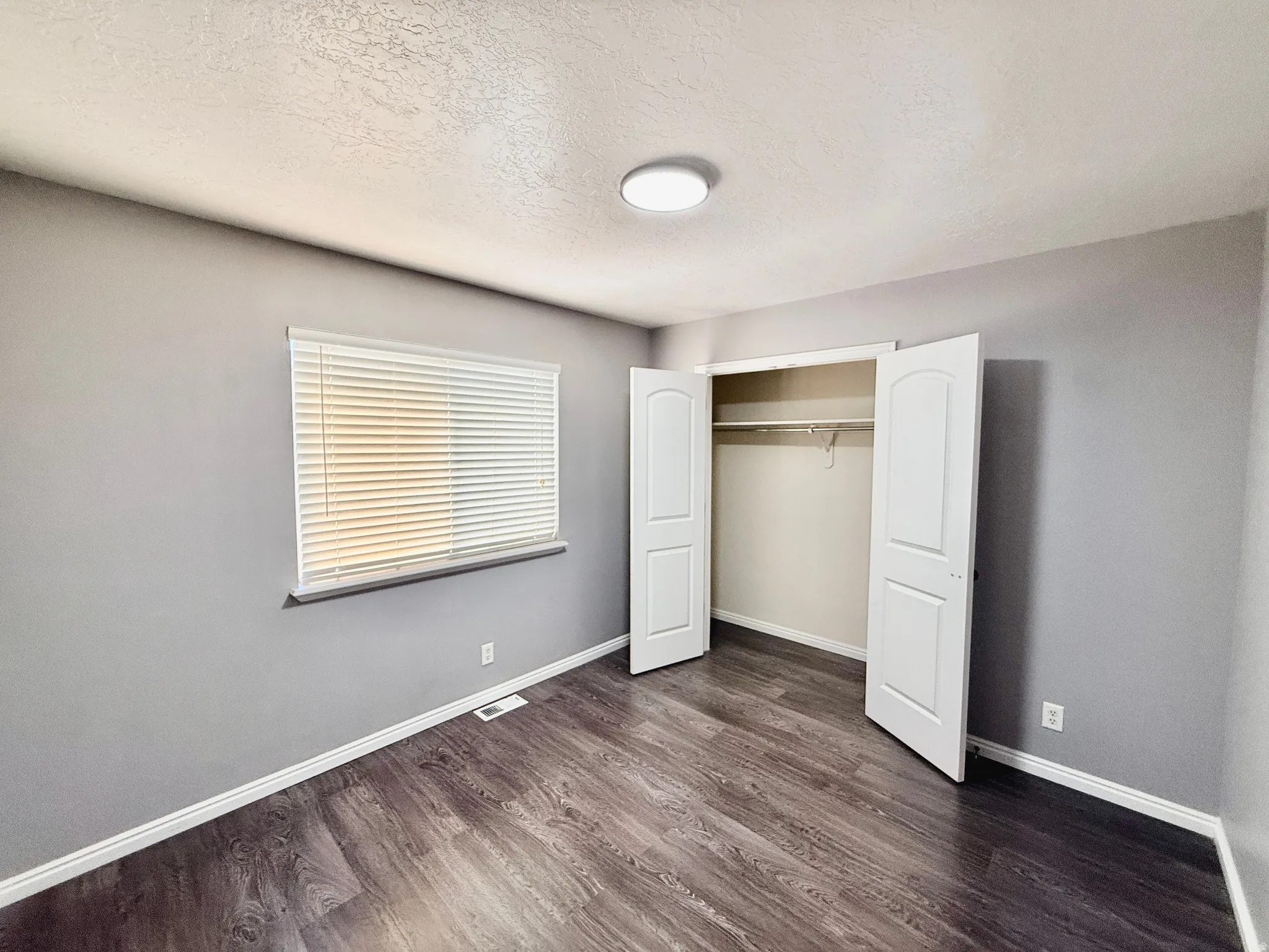 Room featuring wood-finish flooring, a double-door closet with shelving and hanging rod, a window with horizontal blinds, gray painted walls, and a ceiling-mounted light fixture