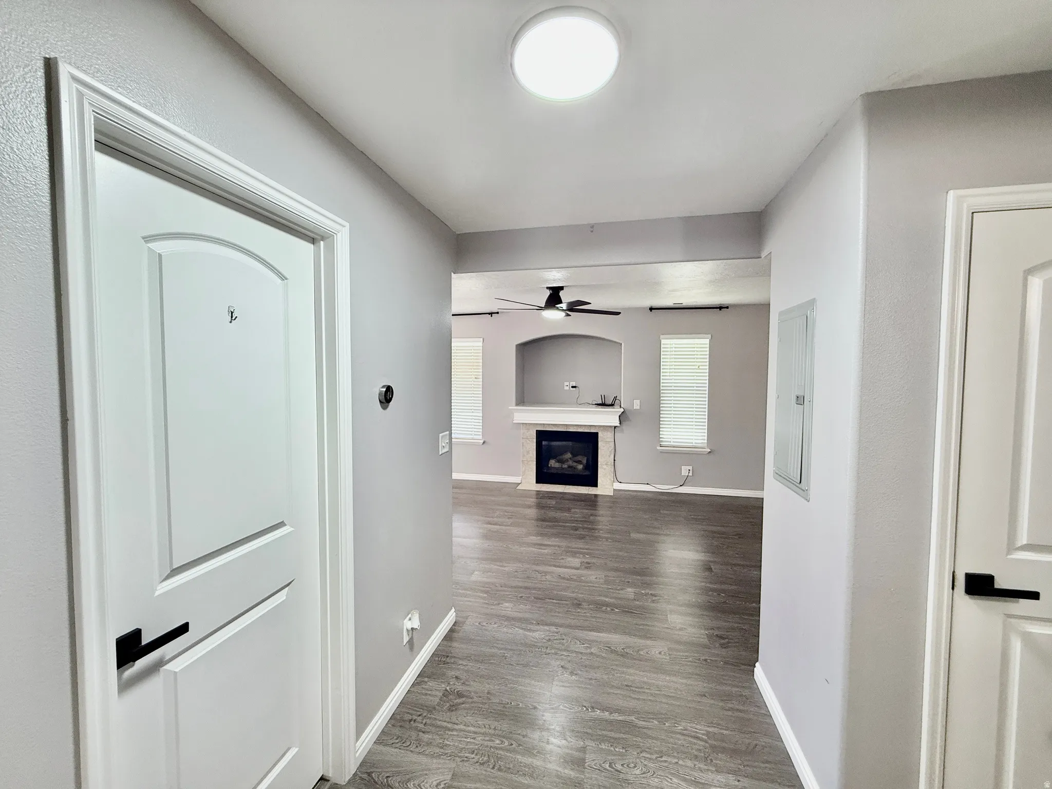 Interior hallway with wood-finish flooring, leading to a living area featuring a fireplace with a light-colored surround, a ceiling fan, and white window blinds