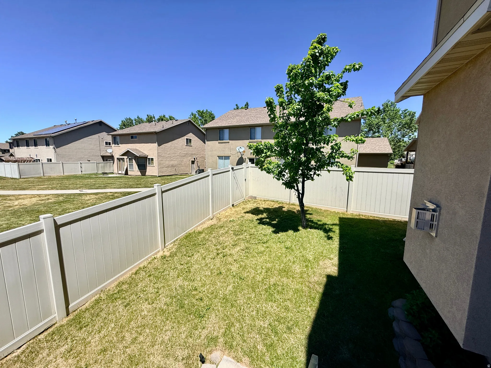 Fenced backyard featuring a mature tree, an exterior wall with a window-mounted air conditioning unit, and a paved area bordering the house