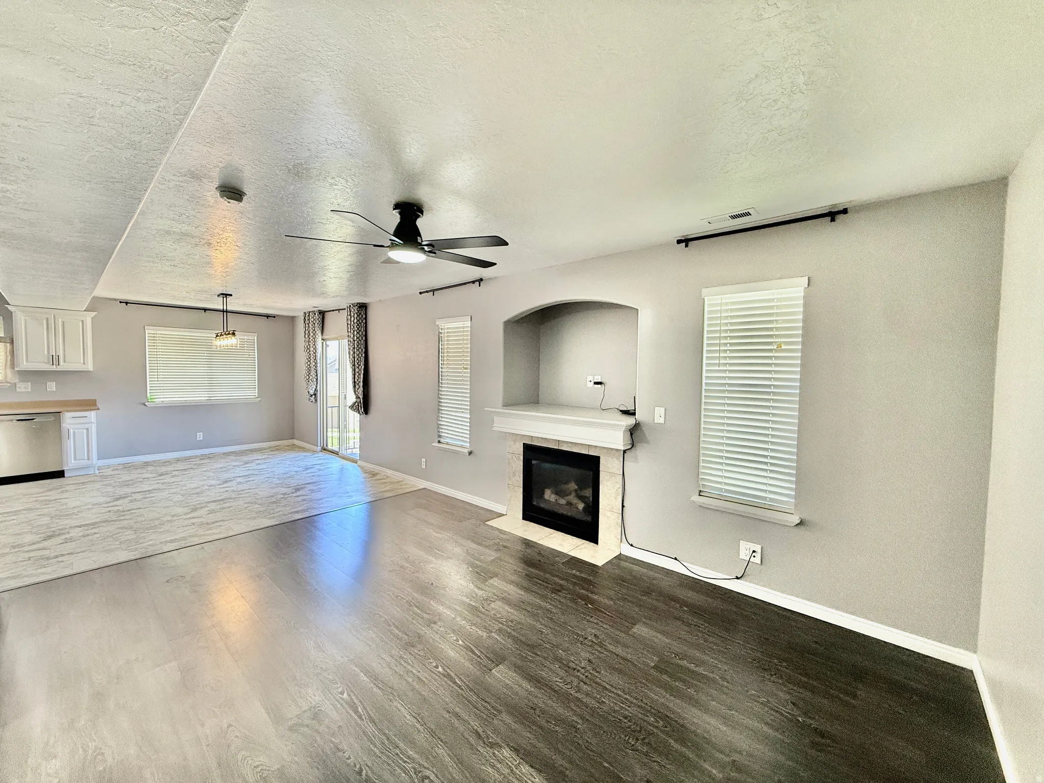 Spacious open-concept living area featuring dark wood-finish flooring, a contemporary ceiling fan, and a built-in fireplace with a mantel and tiled surround