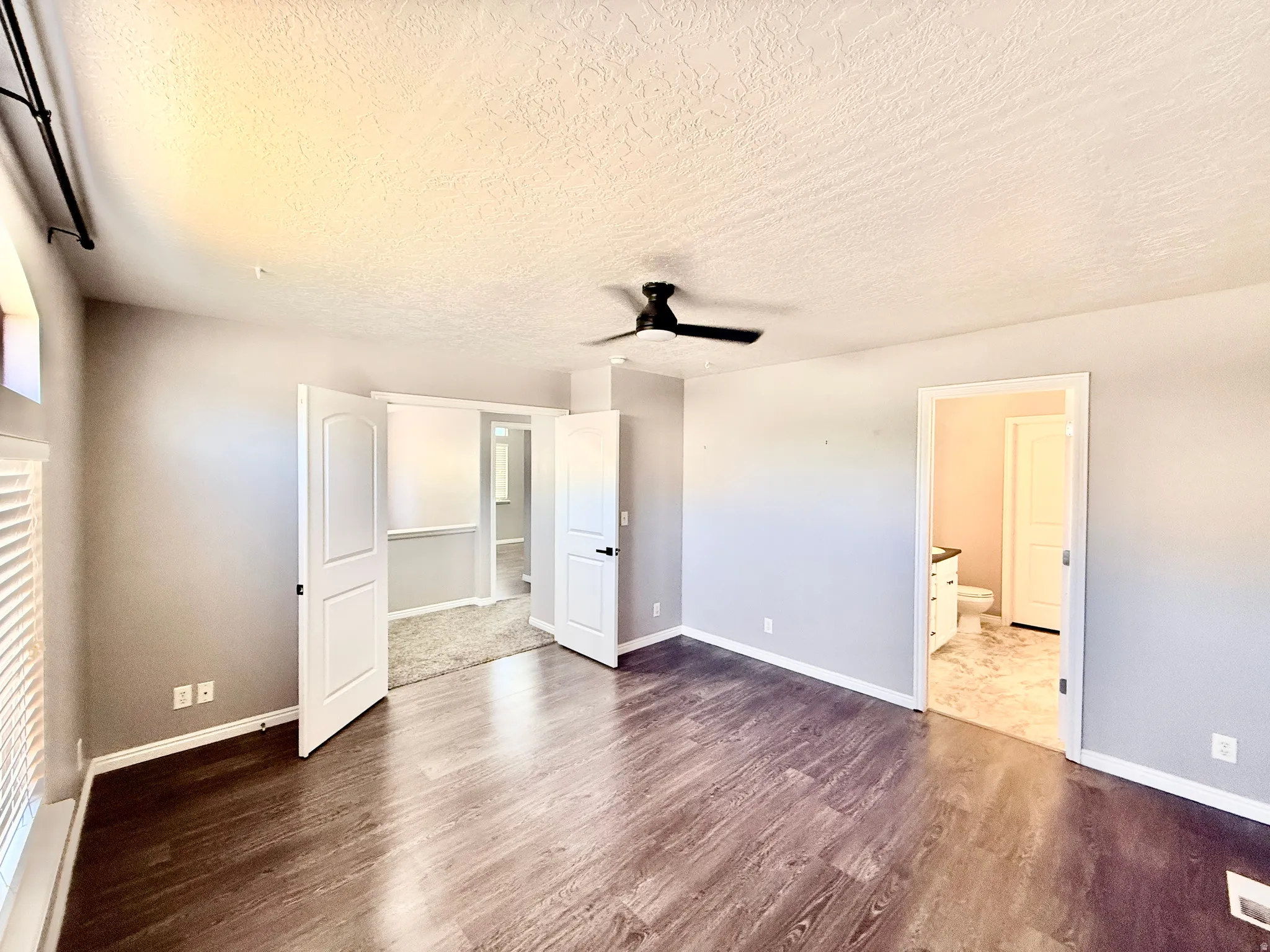 Spacious room featuring wood-finish flooring, a ceiling fan with integrated lighting, window with white louvered blinds, and two paneled doors leading to separate areas