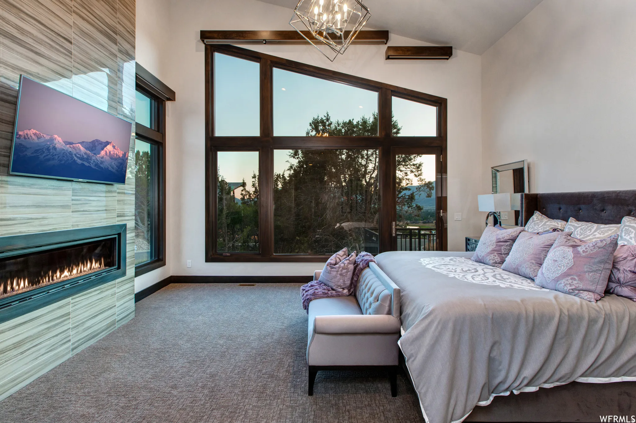 Carpeted bedroom with vaulted ceiling, a tile fireplace, suspended lighting, multiple windows, and access to outside