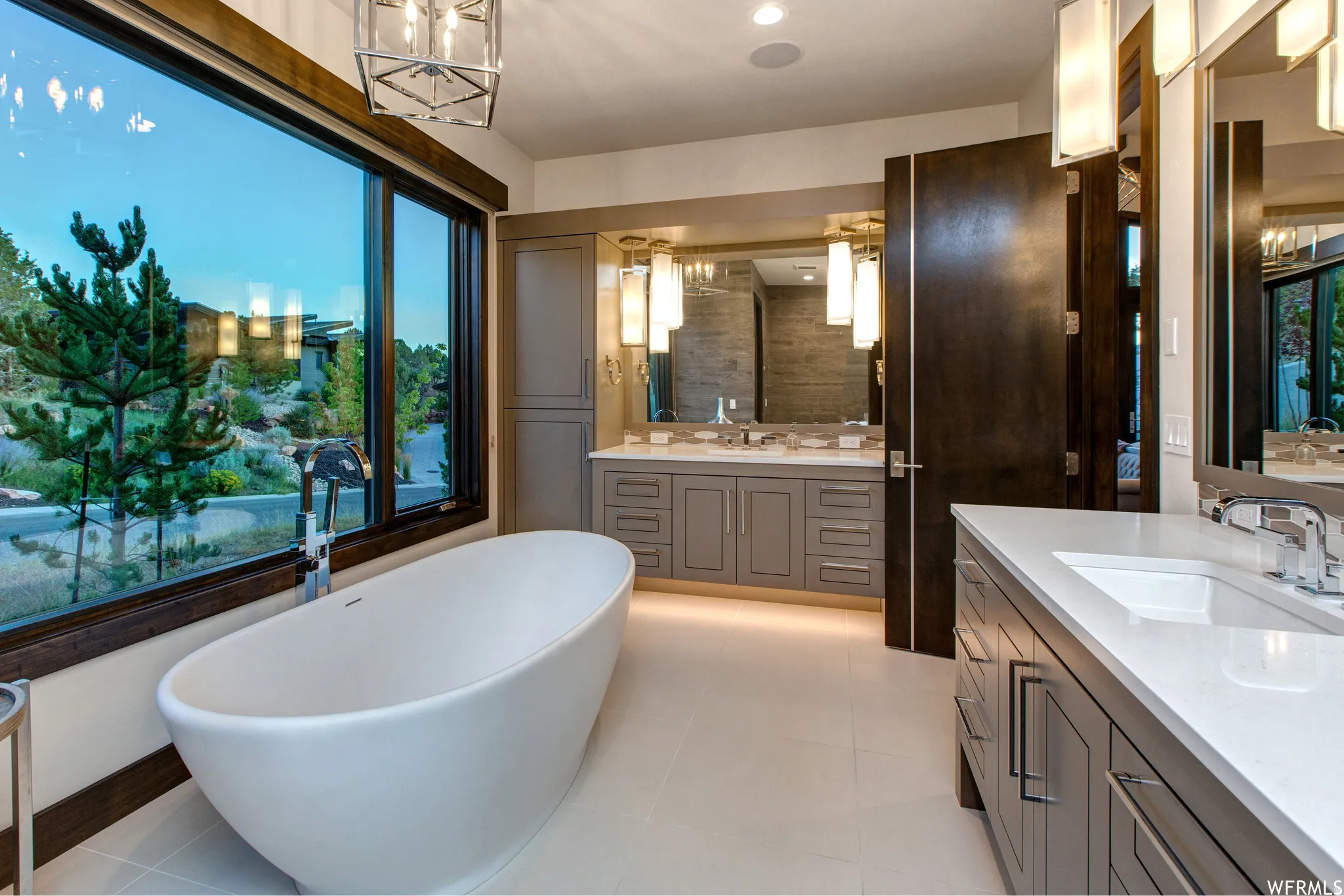 Full bathroom with hanging lights, two vanities, a freestanding tub, and light tile patterned floors