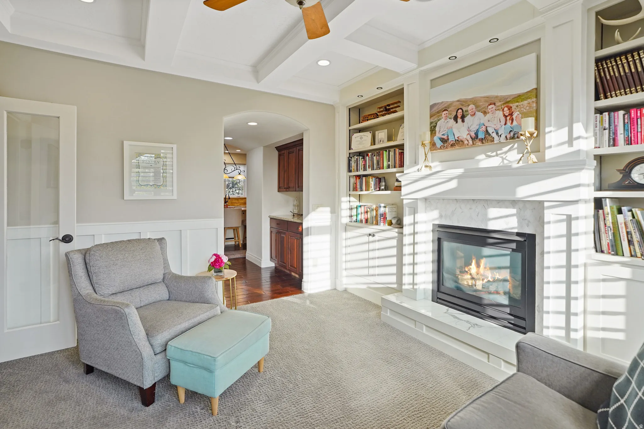 Living area featuring built in features, coffered ceiling, ceiling fan, a glass covered fireplace, and dark colored carpet