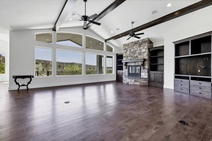 Unfurnished living room with a stone fireplace, built in features, dark wood-type flooring, lofted ceiling, and a ceiling fan