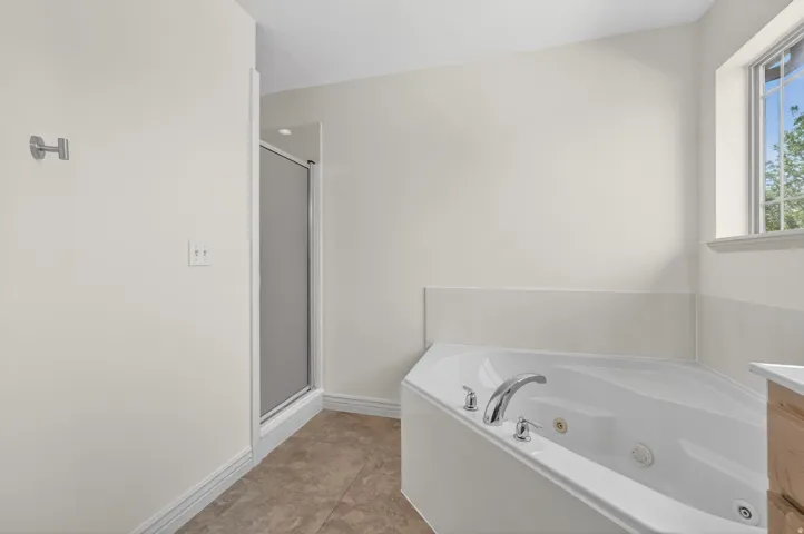 Corner jetted tub with chrome fixtures, enclosed shower, tile flooring, and a window with white trim