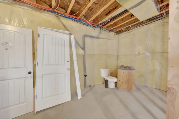 Basement space featuring exposed ceiling joists, insulation with vapor barrier, concrete flooring, and a partial bathroom installation with a toilet and wood-finish vanity