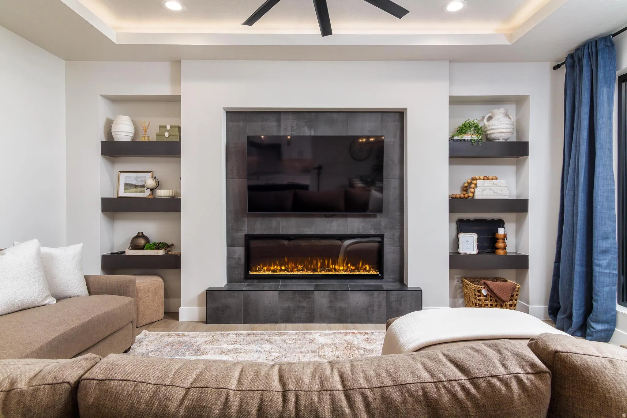 Living area with built in features, a fireplace, wood finished floors, ceiling fan, and a tray ceiling