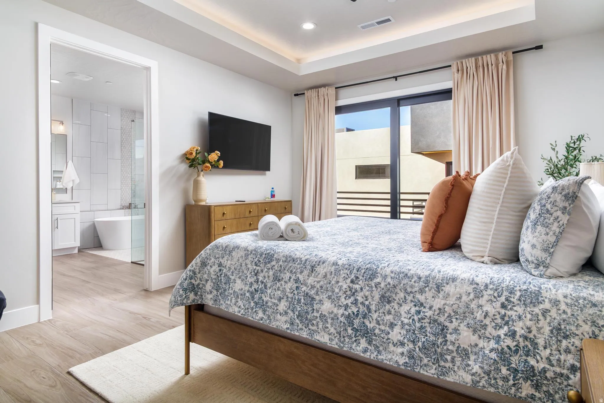 Bedroom with a raised ceiling, access to outside, light wood finished floors, recessed lighting, and ensuite bathroom