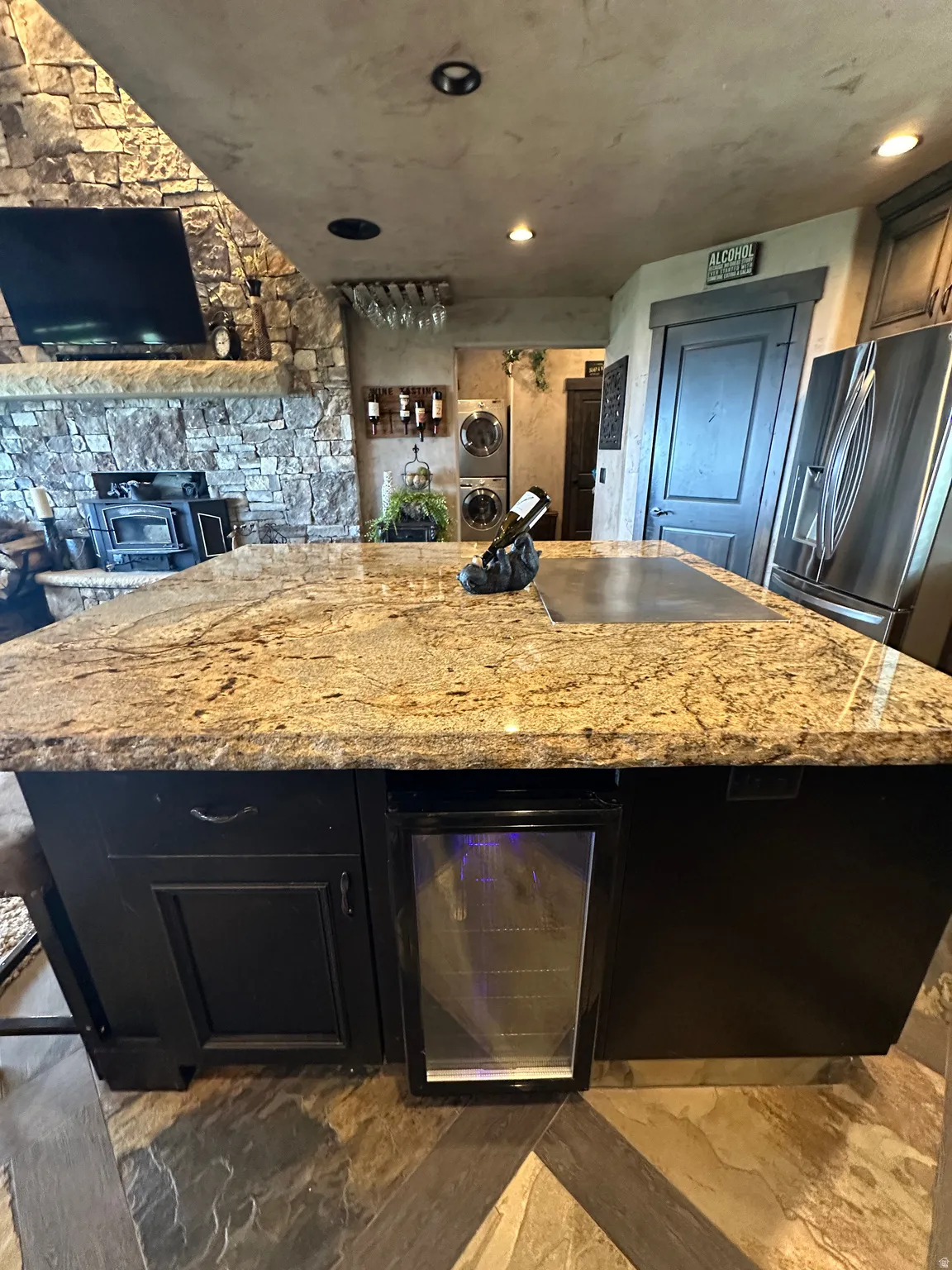 Kitchen with dark cabinetry, a kitchen island, wine cooler, stainless steel fridge with ice dispenser, and light stone countertops
