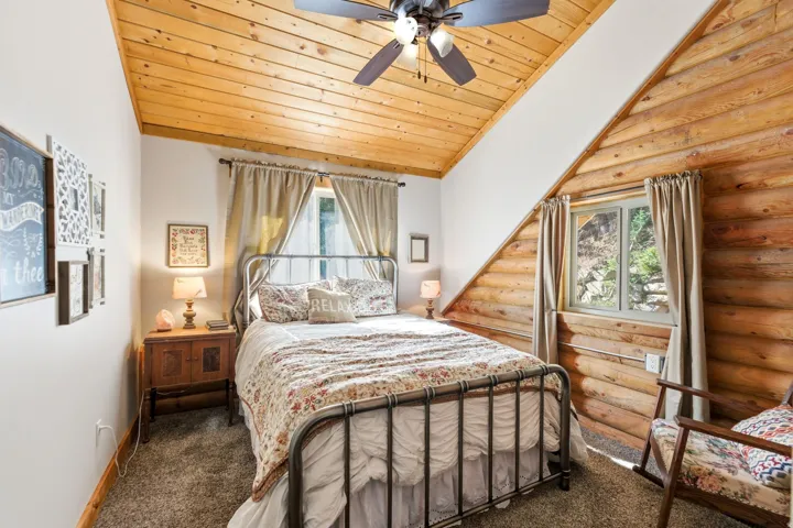 Carpeted bedroom featuring a vaulted wooden ceiling, multiple windows, ceiling fan, and log walls
