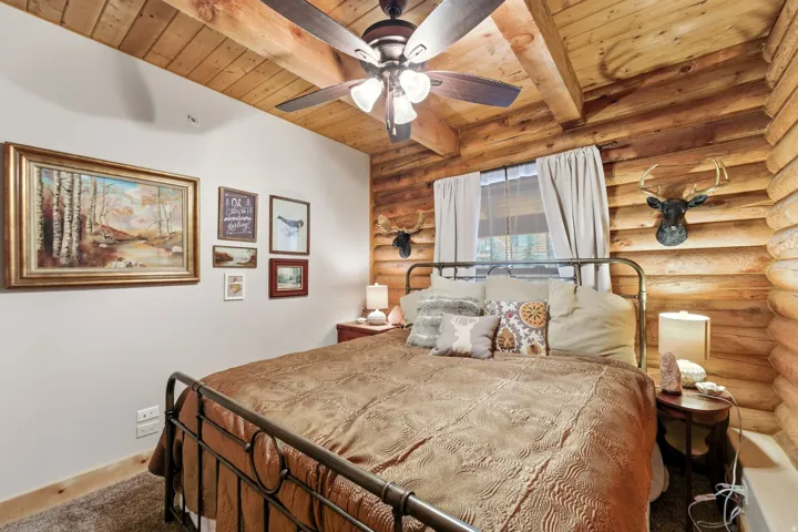 Carpeted bedroom with rustic walls, a ceiling fan, and a wood ceiling with exposed beams