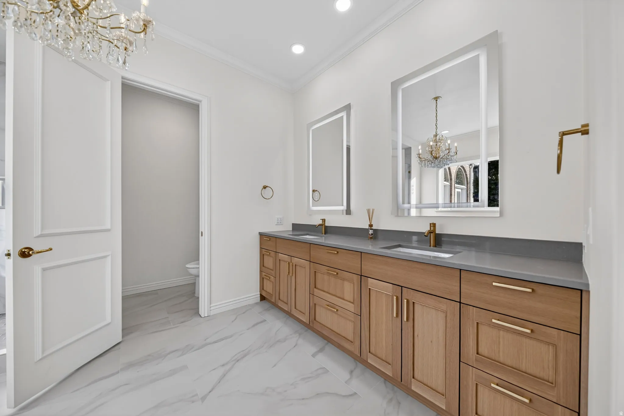 Primary suite bathroom with chandelier, double vanity, light marble finish flooring, and crown molding