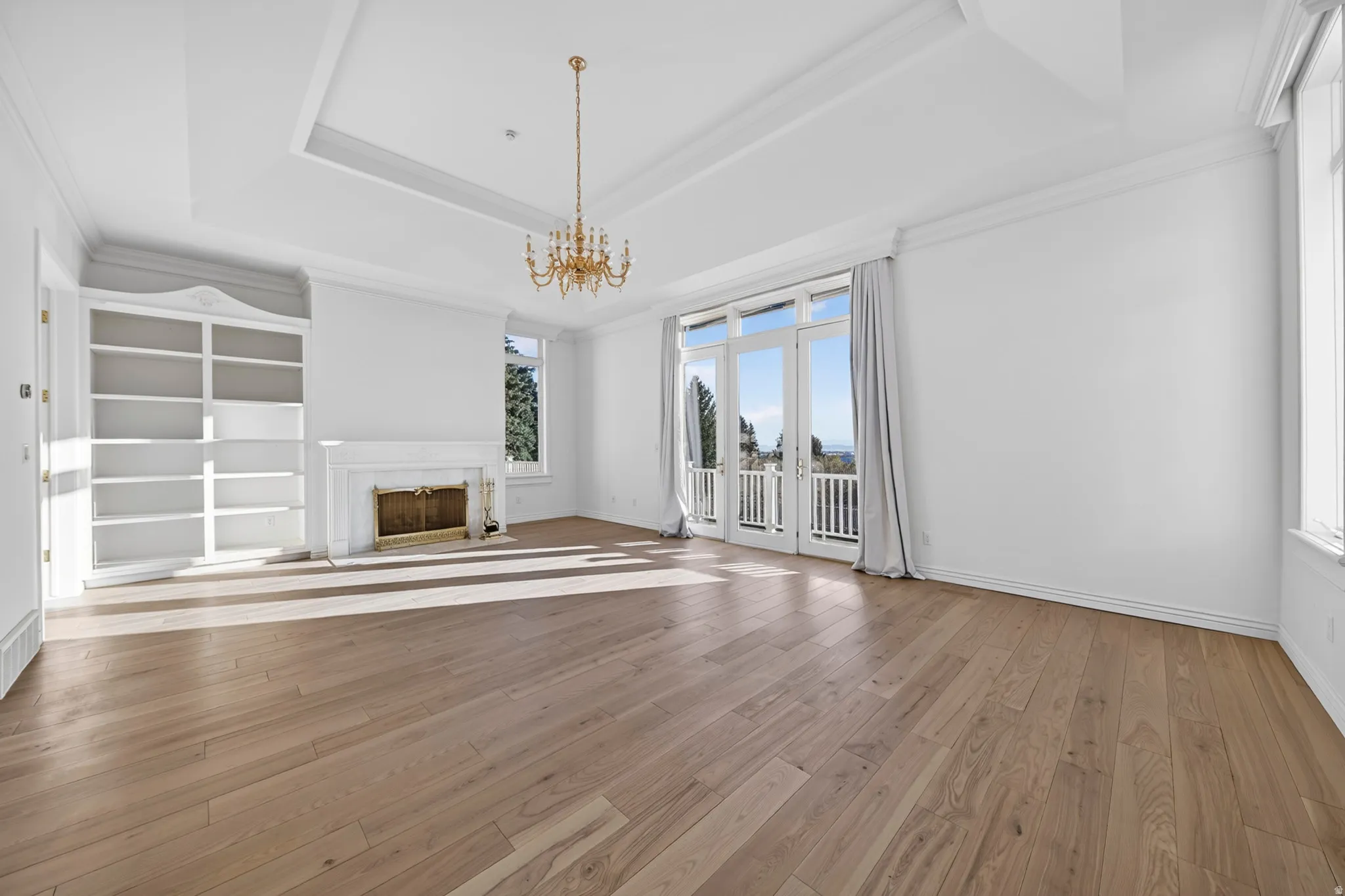 Primary suite with chandelier, a tray ceiling, hardwood flooring, ornamental molding, and a fireplace