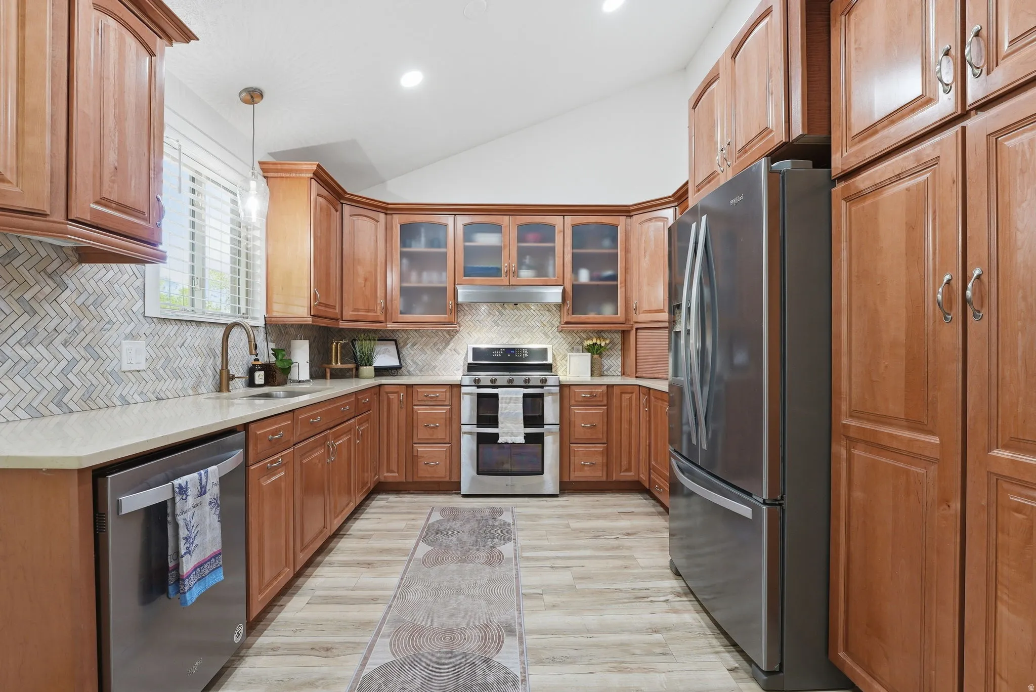 Kitchen featuring vaulted ceiling, stainless steel appliances, wood finish cabinetry, glass fronted cabinets, and hanging light fixtures